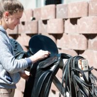 Pretty teenage girl equestrian cleans black Leather Horse Saddle and equipment at farm on bright sunny day. Horizontal outdoors summertime image.